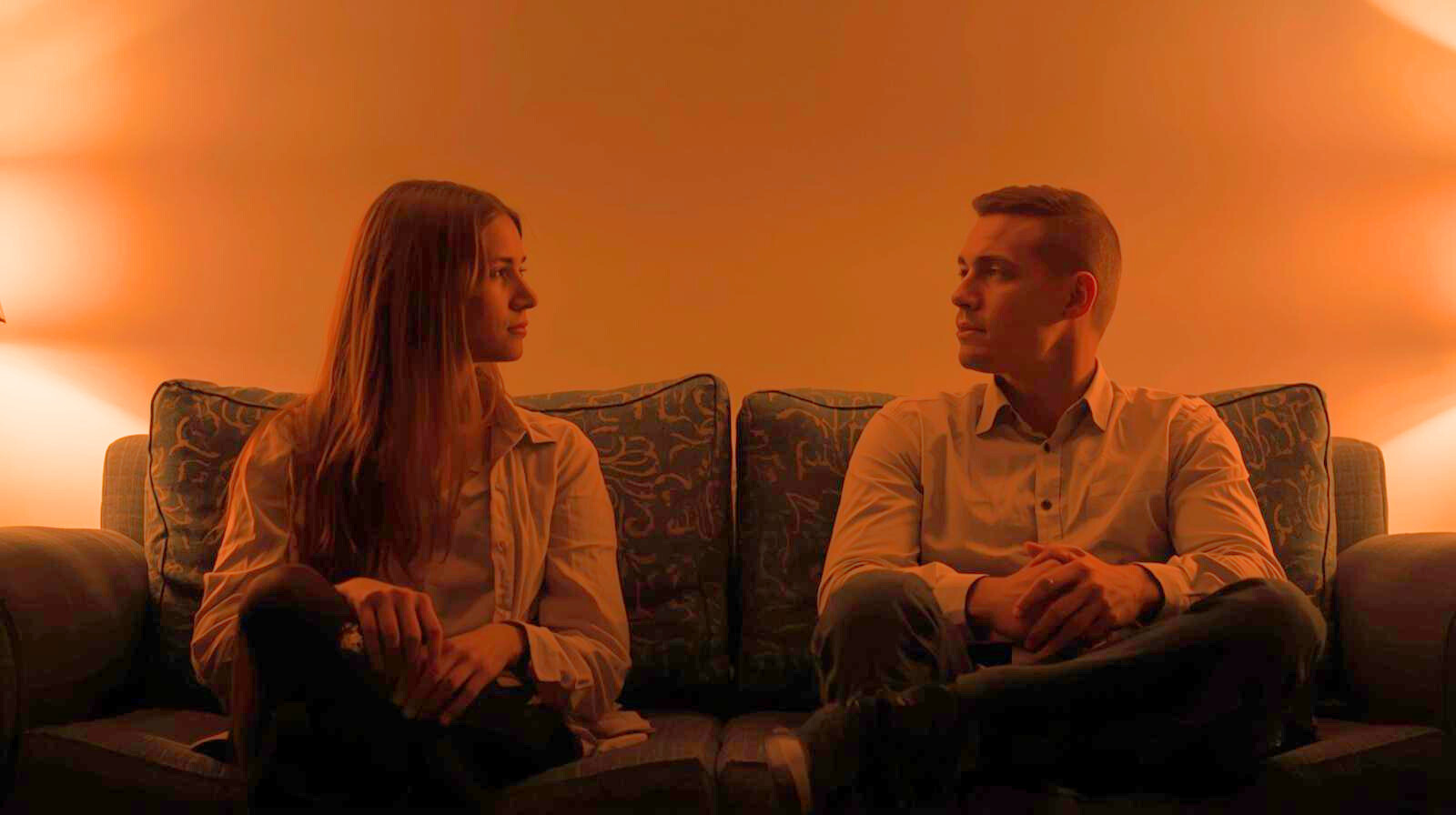 Pareja sentada en extremos opuestos del sofá, con gestos de tensión y desconexión emocional, en una habitación cálida y silenciosa mostrando discusiones constantes